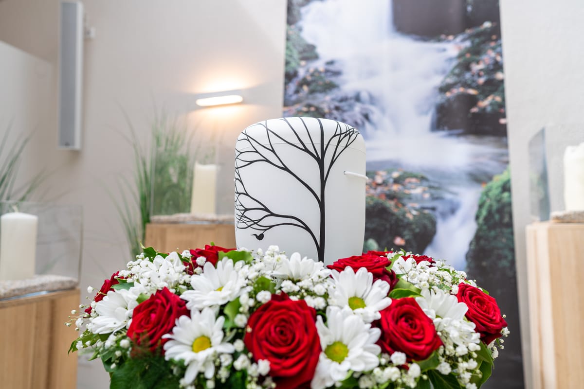 Urne mit Baummotiv, umgeben von roten Rosen und Margeriten