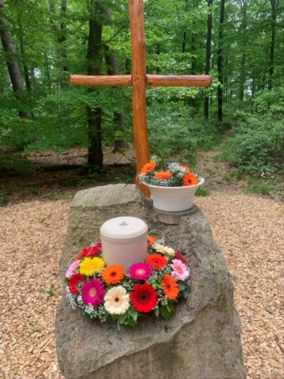 Urne mit Blumenschmuck vor einem Holzkreuz im Wald